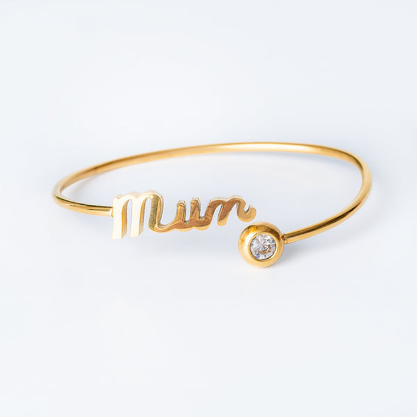 A yellow gold Ghaziza Mum bangle bracelet with the inscription 'Mum' and a heart shape, featuring a small cubic zirconia gemstone.