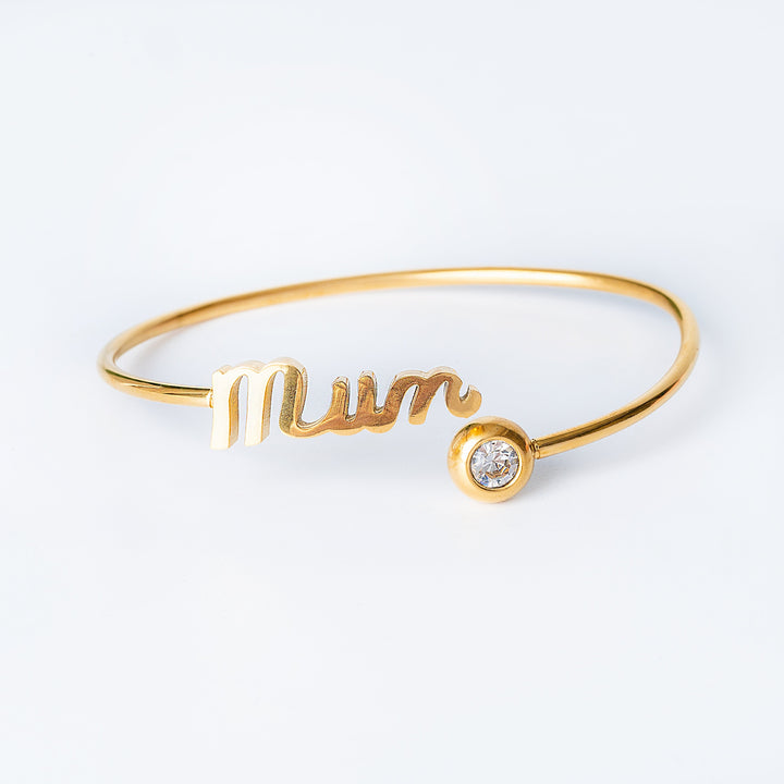 A yellow gold Ghaziza Mum bangle bracelet with the inscription 'Mum' and a heart shape, featuring a small cubic zirconia gemstone.