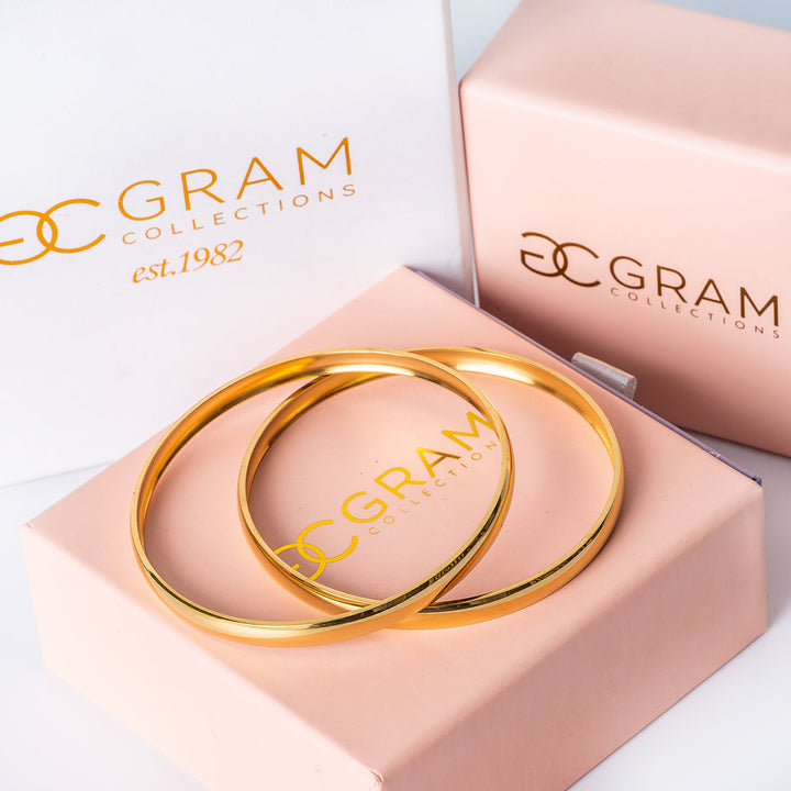 Two gold-colored  Kolina filli stainless steel bracelets with a shiny finish, placed on their uniqly designed pink packages. 