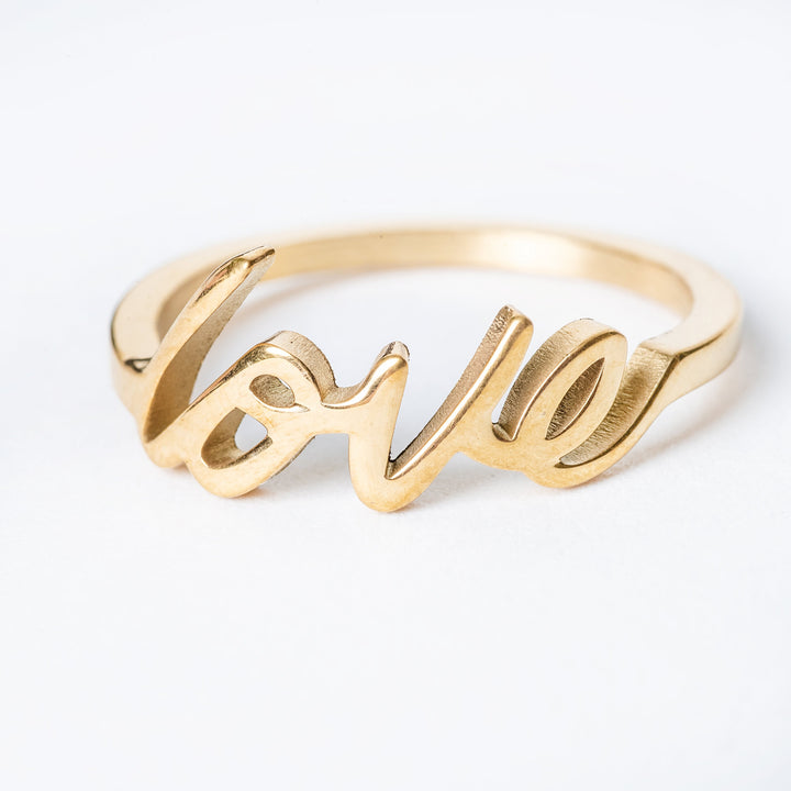 A gold-colored stainless steel love ring with the word 'LOVE' written in cursive on the band.