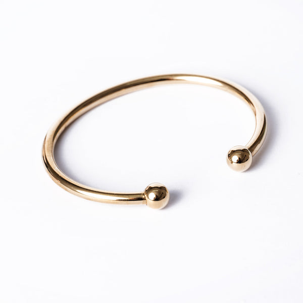 A gold-colored Santi bracelet with a simple design, featuring a circular shape and two spherical ends, displayed against a white background.