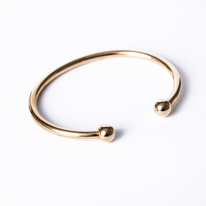A gold-colored Santi bracelet with a simple design, featuring a circular shape and two spherical ends, displayed against a white background.