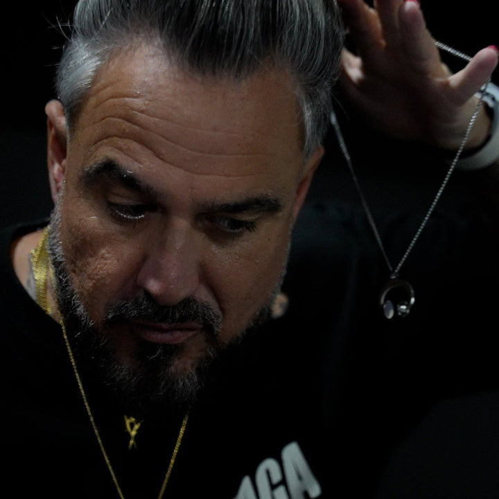 Man with tattoos and a beard wearing a black hoodie with visible text, while holding dj necklace in his nands.
