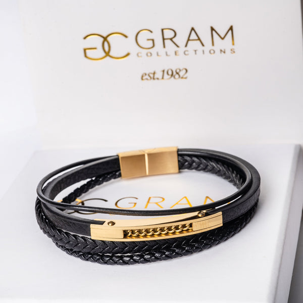A black leather Andrea Gold bracelet with yellow gold plating, presented on a white box with 'GRAM Collections' branding.