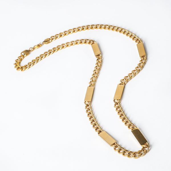 A gold-colored stainless steel Maltese style bar necklace laid out on a white surface.