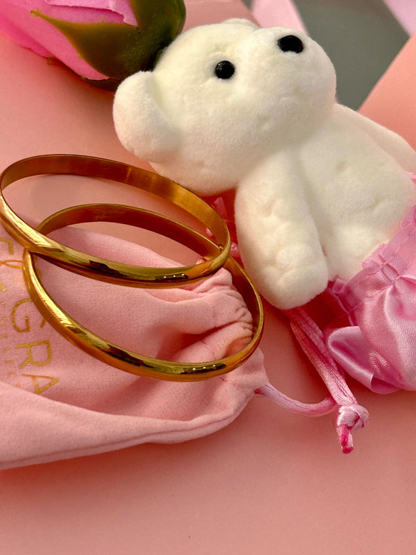 A pair of yellow gold-plated stainless steel Fili bracelets displayed on a pink surface, with a white plush toy and pink tissue paper nearby.