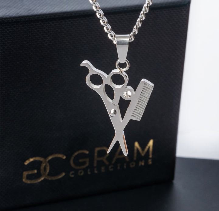 The gram collections hairdresser necklace standing in the center of the image against a white background, and in front of its uniquely designed box. 
