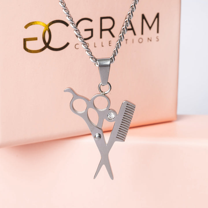 a gram collections The Hairdresser Necklace standing in the center of the image, in front of its uniquely designed pink boxes, against a white background. 
