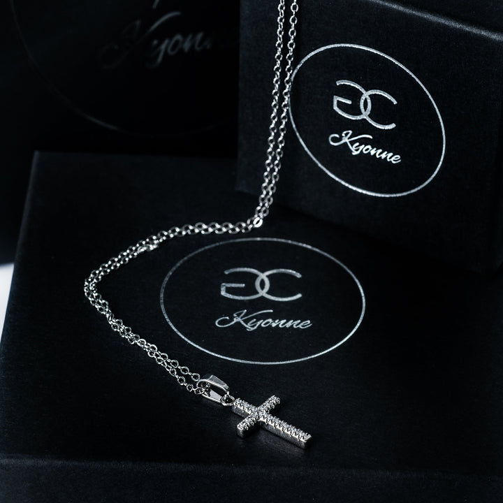 Silver Kyonne cross necklace with clear diamonds displayed on black branded boxes