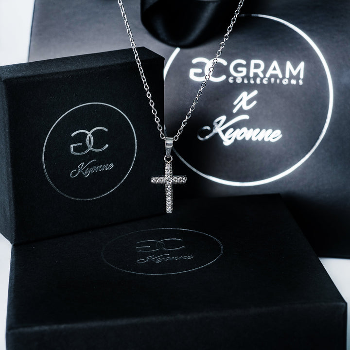 Silver Kyonne cross necklace with clear diamonds displayed on black branded boxes