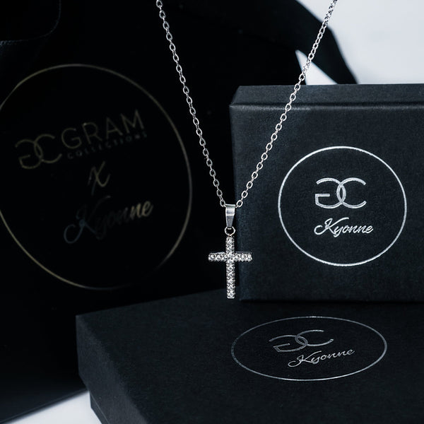 Silver Kyonne cross necklace with clear diamonds displayed on black branded boxes