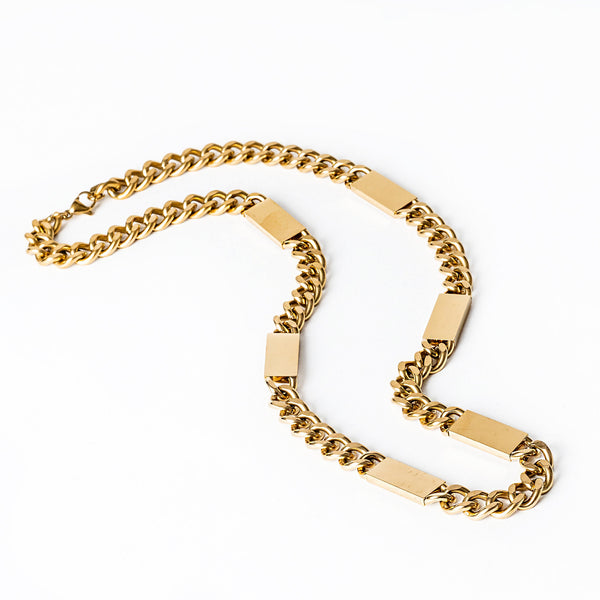 A gold-colored stainless steel Large bar necklace with a curb chain design laid out on a white surface.
