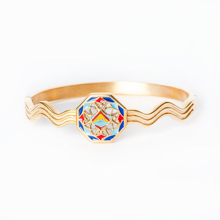 A close up view of gold Luzzu bracelet with a colorful hexagonal emblem on a white background