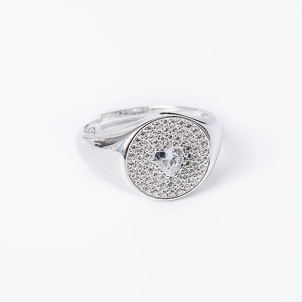 A silver Rena ring with a round face, featuring a central cubic zirconia gemstone, surrounded by smaller clear gems in a heart-shaped pattern.
