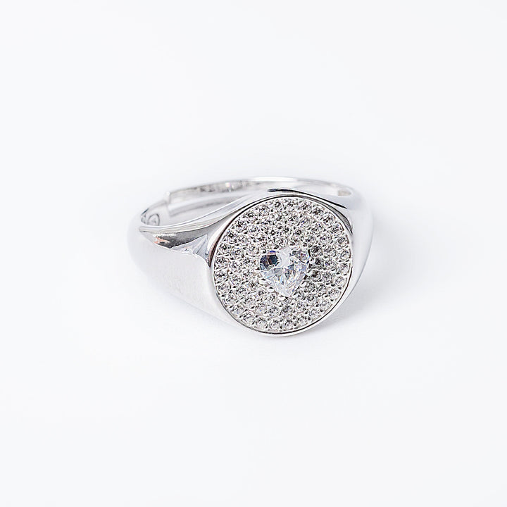 A silver Rena ring with a round face, featuring a central cubic zirconia gemstone, surrounded by smaller clear gems in a heart-shaped pattern.