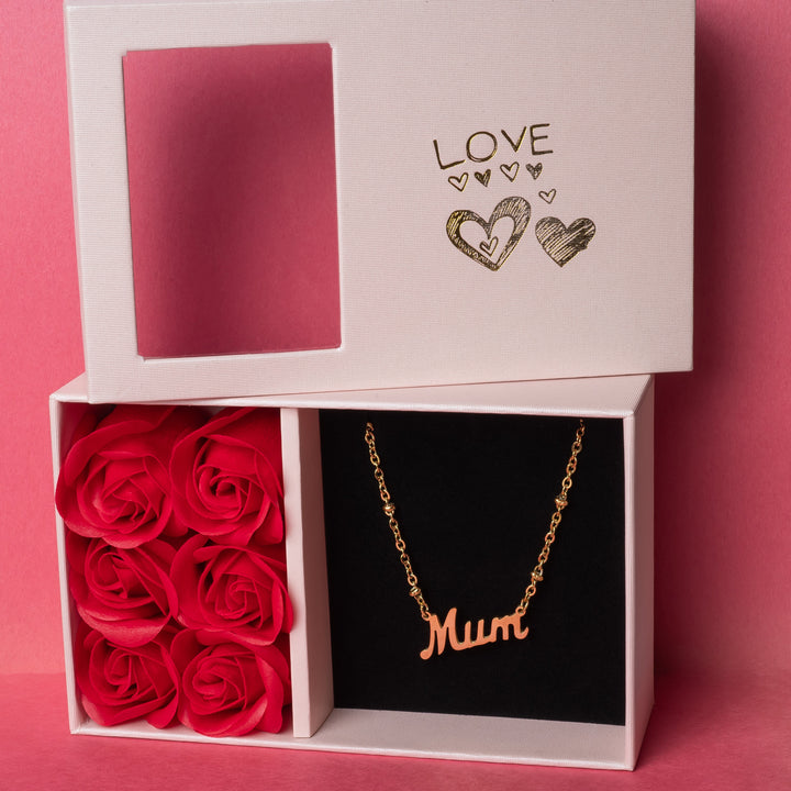 Rosina mum necklace with a gold-plated pendant, engraved with the word 'Mum', presented in a white box with 'LOVE' and hearts design, accompanied by a doily and red roses.