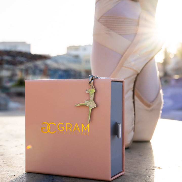 The Ballerina Necklace placed on the edge of its pink box, standing in front of balerina's feet. 