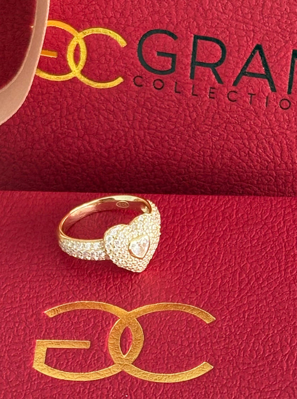 A gold-colored ring with a heart-shaped design, featuring cubic zirconia, displayed on a red surface with the 'QALBI' logo.