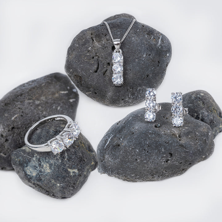 ATrilogy Earrings set, consist of pendant, and ring, each placed on a different dark grey rock, standing in the center of the image against a white background. 