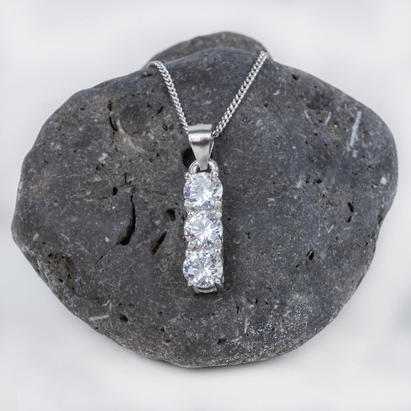 A silver Trilogy necklace with a single AAA cubic zirconia gemstone pendant set in a silver cap, displayed on a textured gray rock.