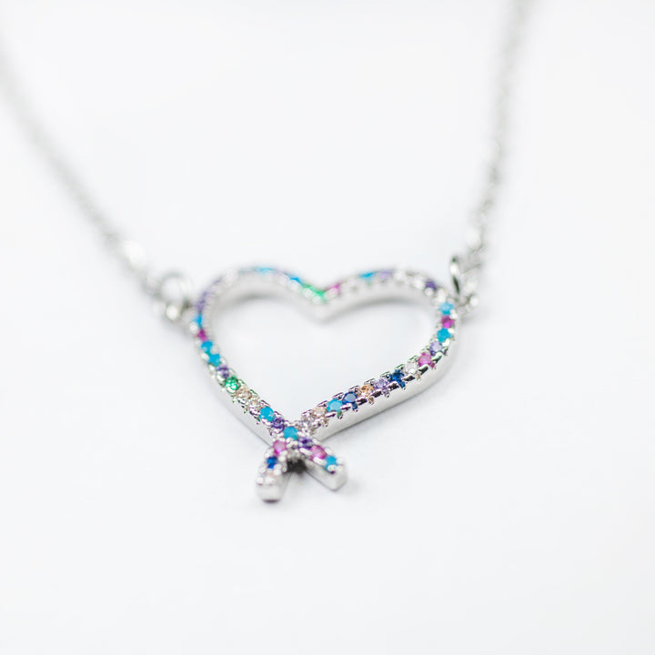 A silver-colored stainless steel necklace featuring a heart-shaped pendant adorned with multicolored cubic zirconia gems. The pendant has a reflective surface and is attached to a silver chain.