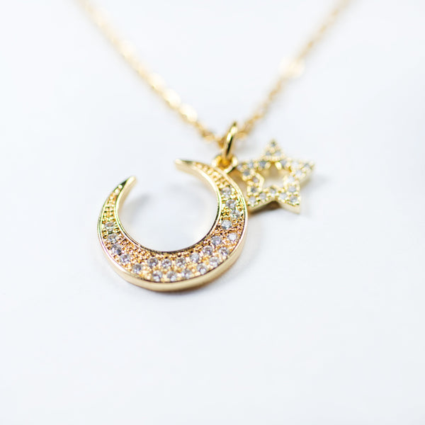 A gold-plated stainless steel pendant in the shape of a crescent moon with a star, encrusted with cubic zirconia, on a white chain.