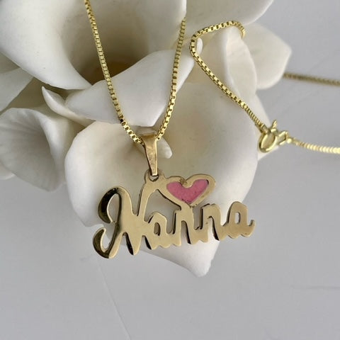 A yellow gold necklace with a heart-shaped pendant featuring the name 'Nanna' and a pink smalt bead, displayed on a white flower-like object.