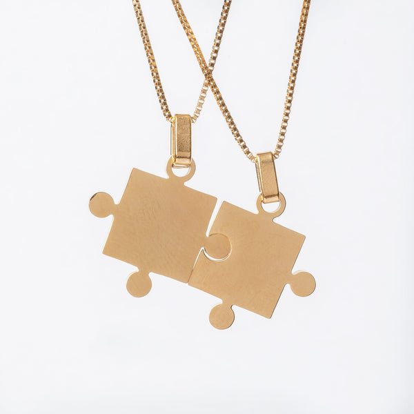 Two gold-colored stainless steel puzzle piece necklaces connected and laid next to each other on a white background.