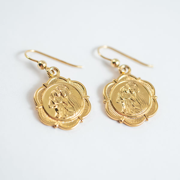 A pair of gold-colored hoop earrings with a floral pattern and a central figure motif on a white background, displayed against a white background.