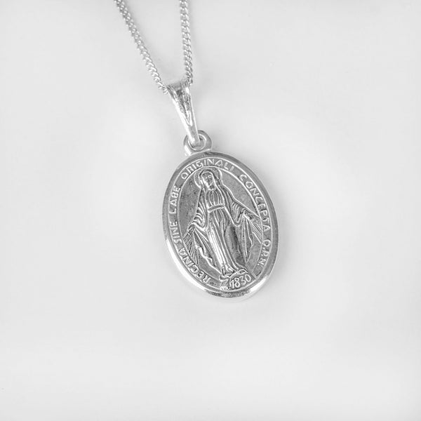 A close up view of Silver Holy Mary Charm Pendant standing in the center of the image against a silver background. 
